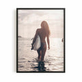 Surfer girl walking into the ocean at golden hour carrying a surfboard during sunset, creating a peaceful coastal photography framed wall art print.