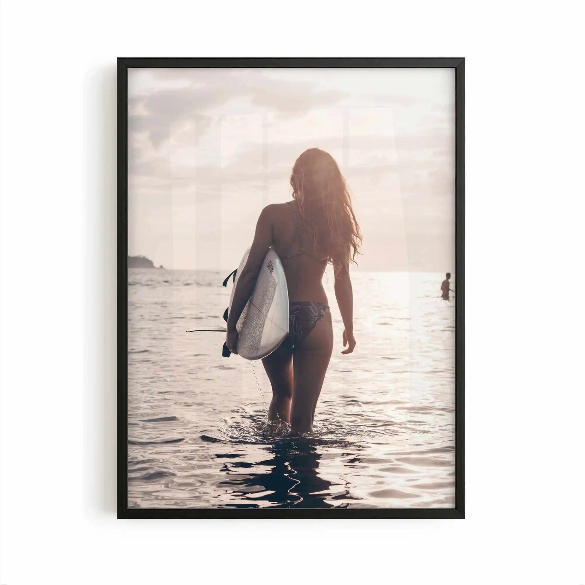 Surfer girl walking into the ocean at golden hour carrying a surfboard during sunset, creating a peaceful coastal photography framed wall art print.