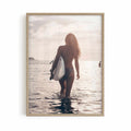 Surfer girl walking into the ocean at golden hour carrying a surfboard during sunset, creating a peaceful coastal photography framed wall art print.