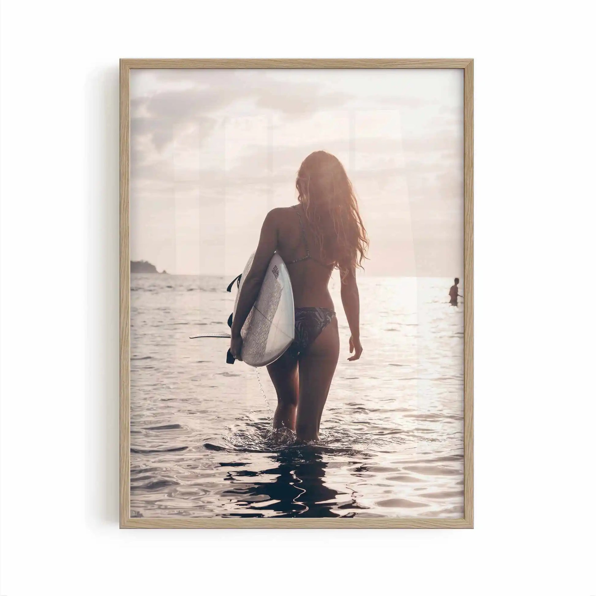Surfer girl walking into the ocean at golden hour carrying a surfboard during sunset, creating a peaceful coastal photography framed wall art print.